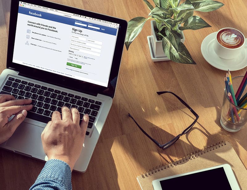 Man working on facebook and signing in to his account with a full desk setup, glasses, pencils, coffee, pot plant, while typing