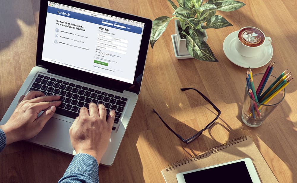 Man working on facebook and signing in to his account with a full desk setup, glasses, pencils, coffee, pot plant, while typing