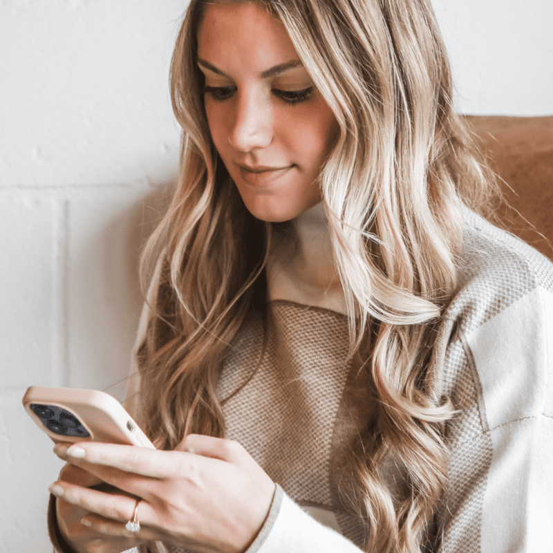 Woman with long blonde hair sitting in a cozy chair, looking down and smiling while using her smartphone.