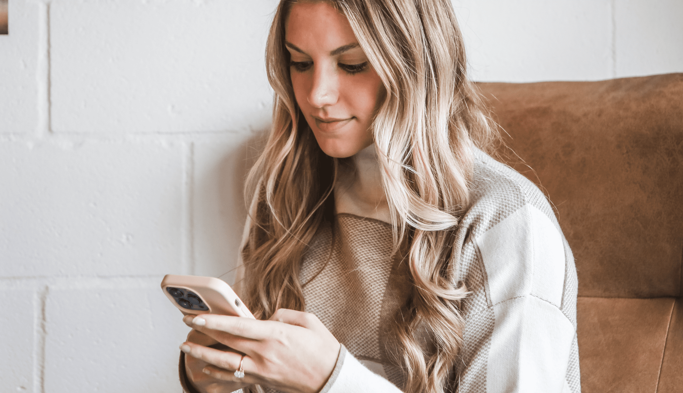 Woman with long blonde hair sitting in a cozy chair, looking down and smiling while using her smartphone.