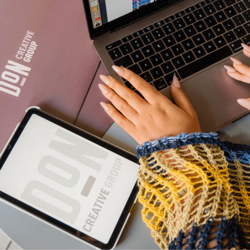 Hands with manicured nails typing on a MacBook beside a Don Creative Group folder and tablet on a round table.