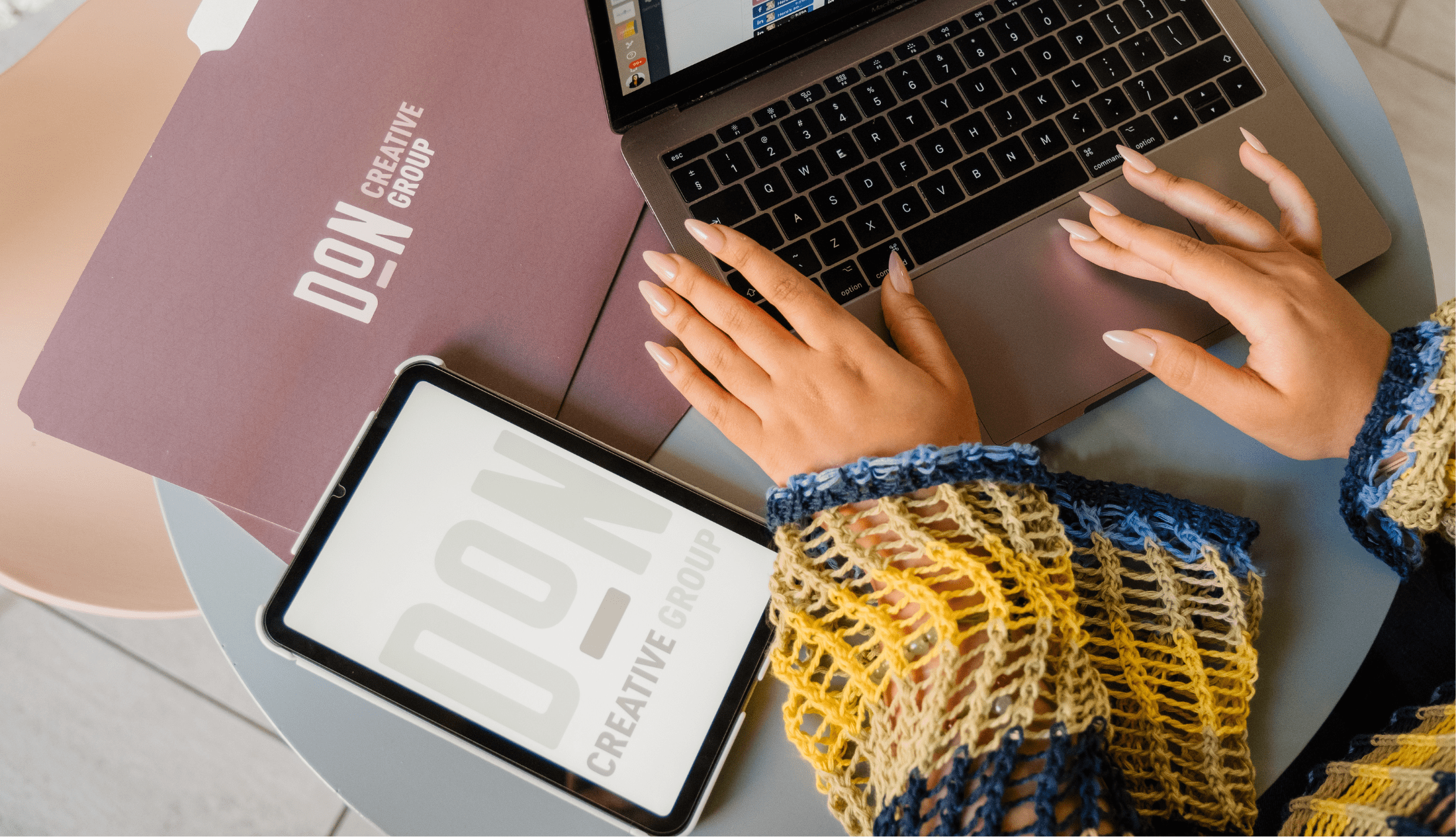 Hands with manicured nails typing on a MacBook beside a Don Creative Group folder and tablet on a round table.