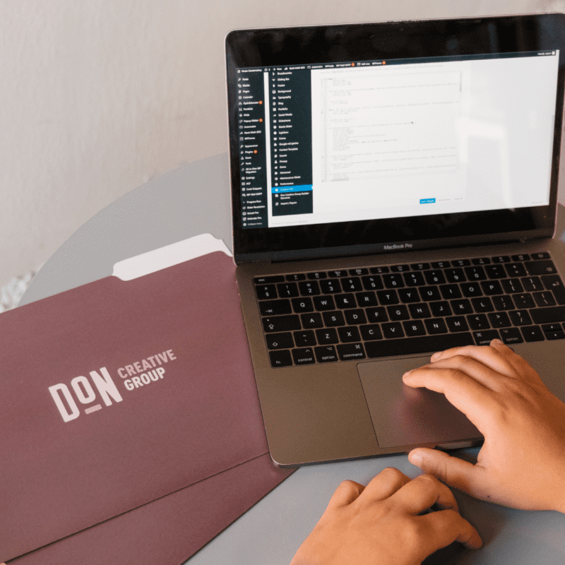 Hands on a laptop editing a WordPress website, with Don Creative Group branded folders nearby on a round table.