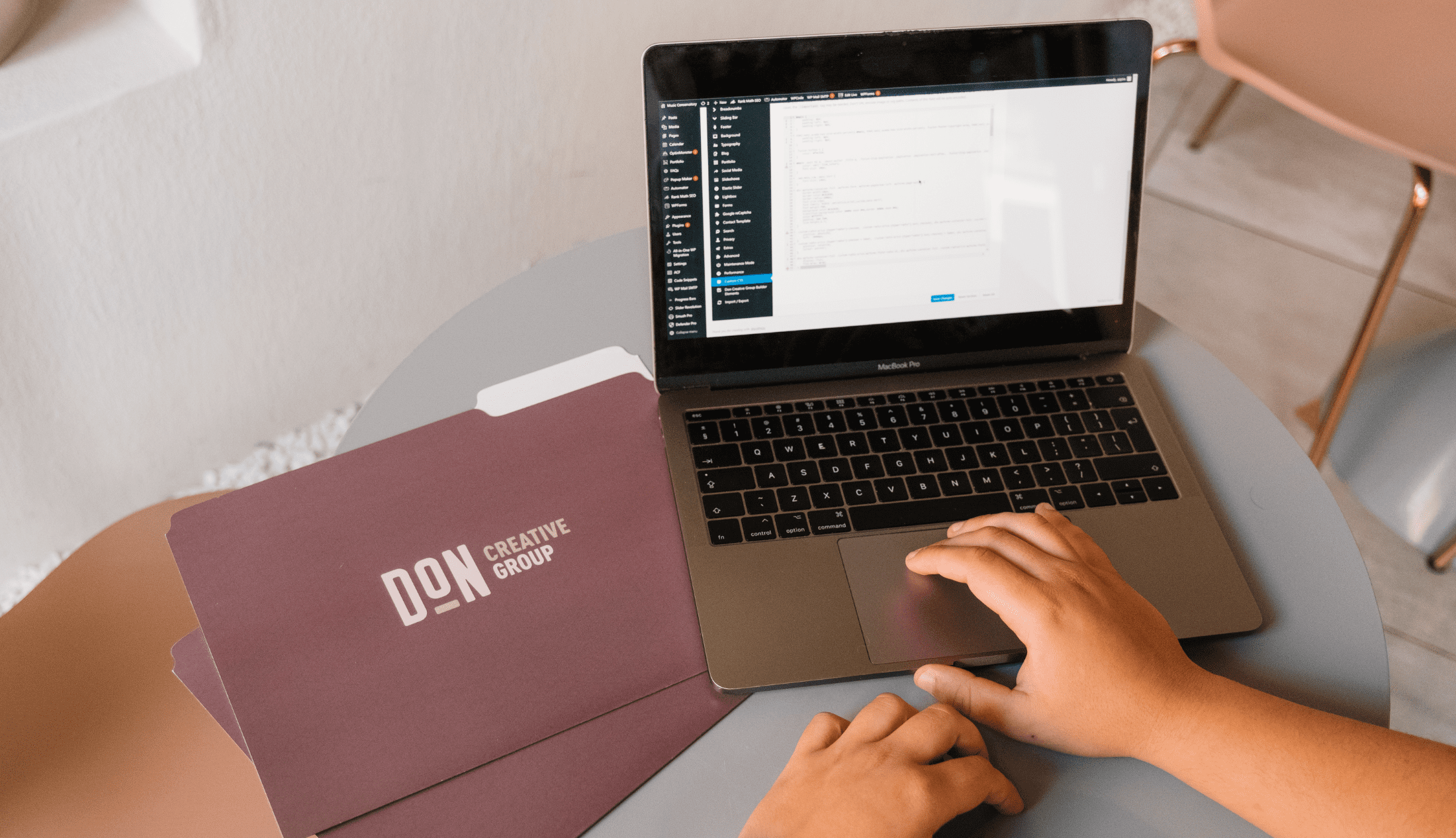 Hands on a laptop editing a WordPress website, with Don Creative Group branded folders nearby on a round table.