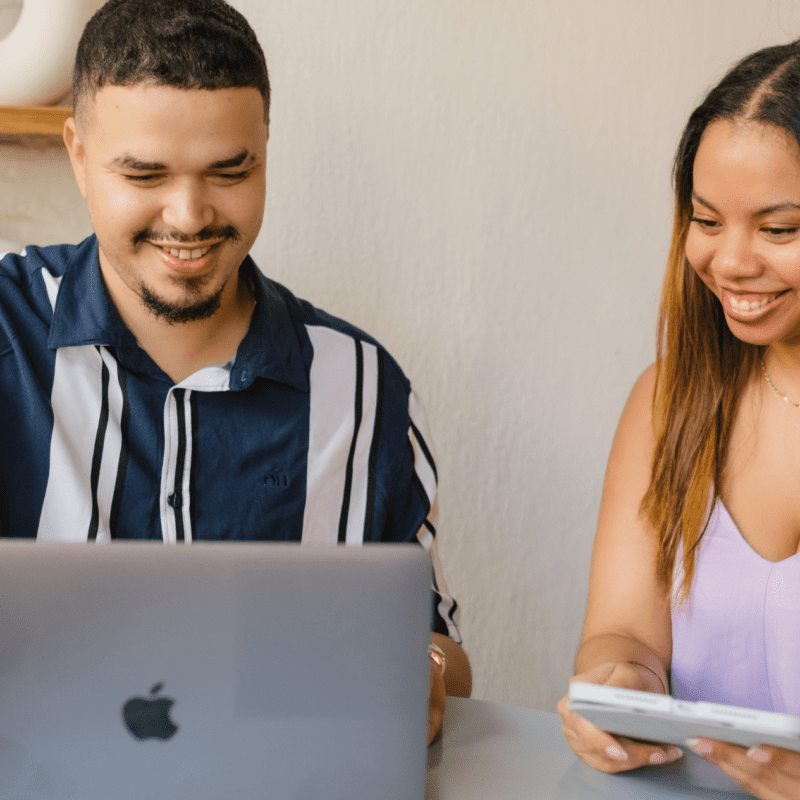 Two professionals smiling while reviewing digital content on a laptop and smartphone together