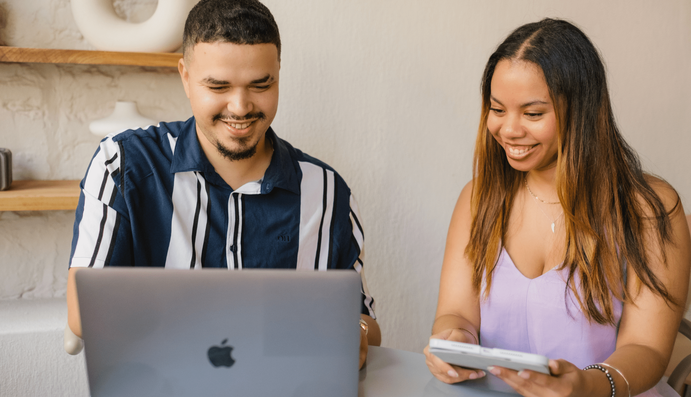 Two professionals smiling while reviewing digital content on a laptop and smartphone together