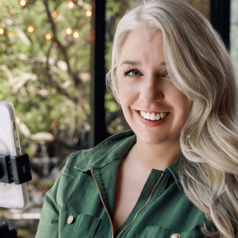 Smiling woman with long blonde hair filming a video using a smartphone on a tripod, in a bright room with natural light and greenery outside.
