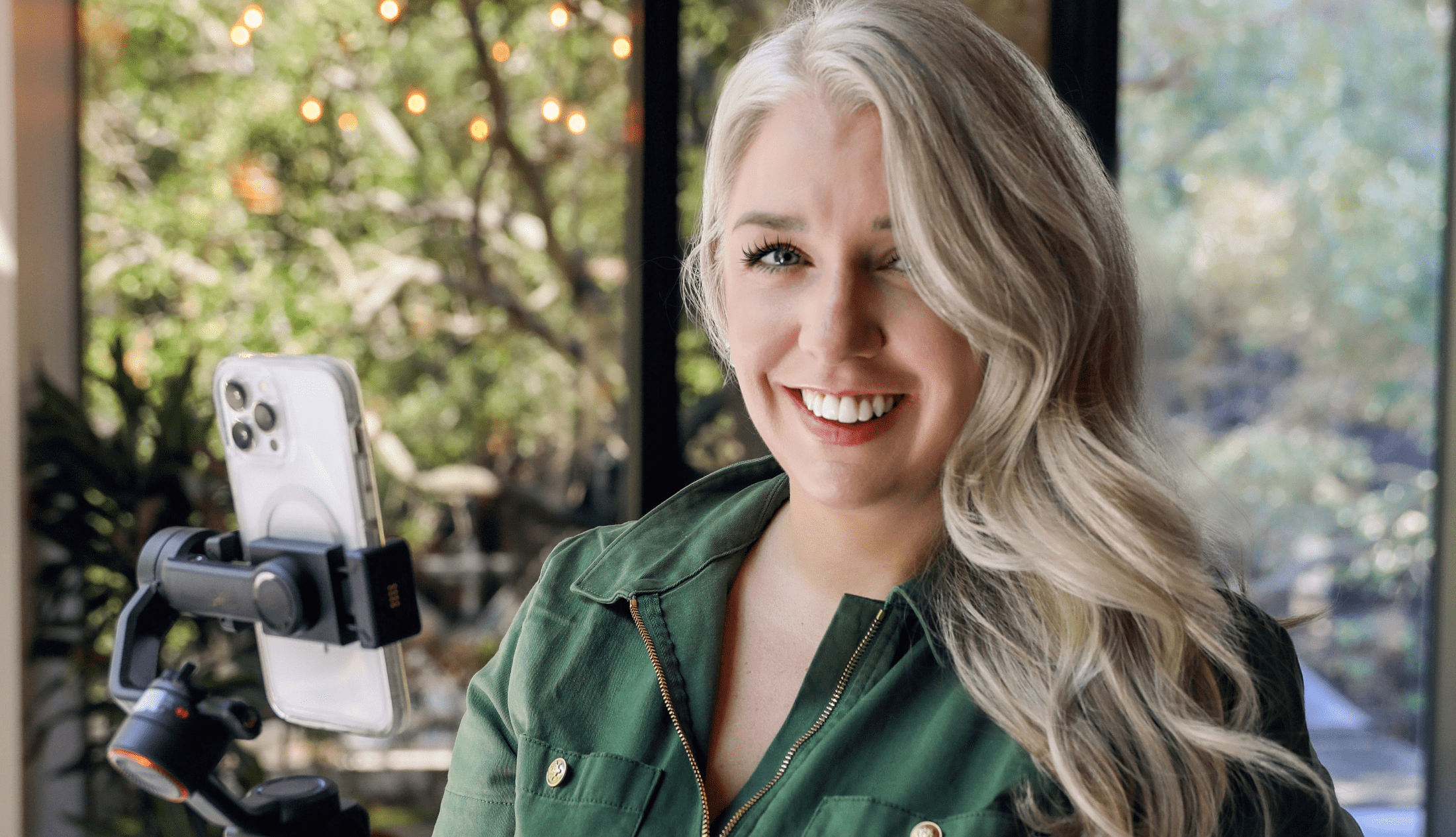 Smiling woman with long blonde hair filming a video using a smartphone on a tripod, in a bright room with natural light and greenery outside.
