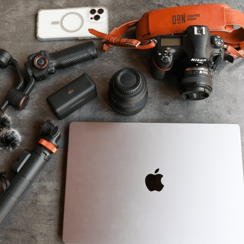 Flat lay of professional content creation gear including a Nikon camera with Don Creative Group strap, lenses, smartphone, gimbal, tripod, and laptop.
