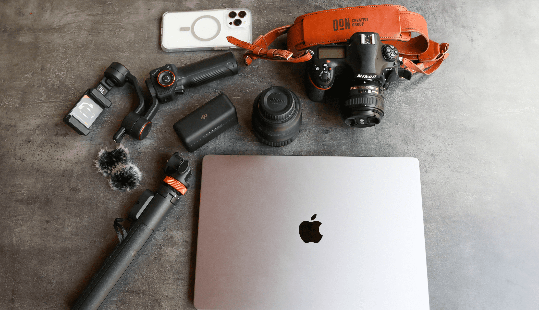 Flat lay of professional content creation gear including a Nikon camera with Don Creative Group strap, lenses, smartphone, gimbal, tripod, and laptop.