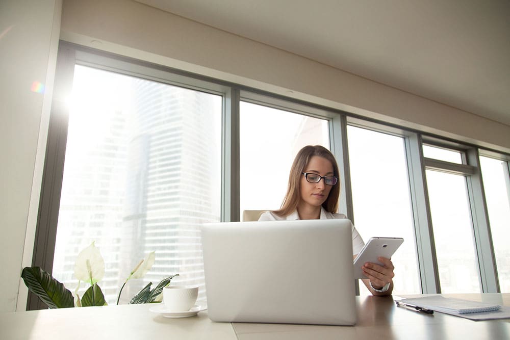 Professional woman in a modern office using a tablet and laptop while working at a desk with a city skyline in the background.
