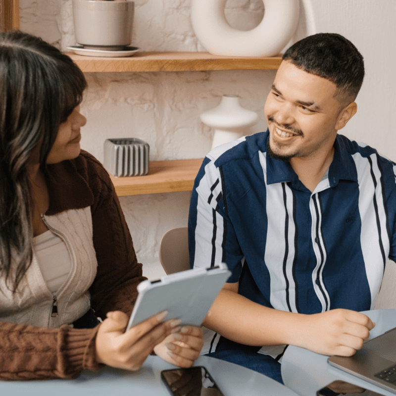 Two team members collaborating at a table with a laptop and tablet, smiling during a creative strategy session in a modern workspace.