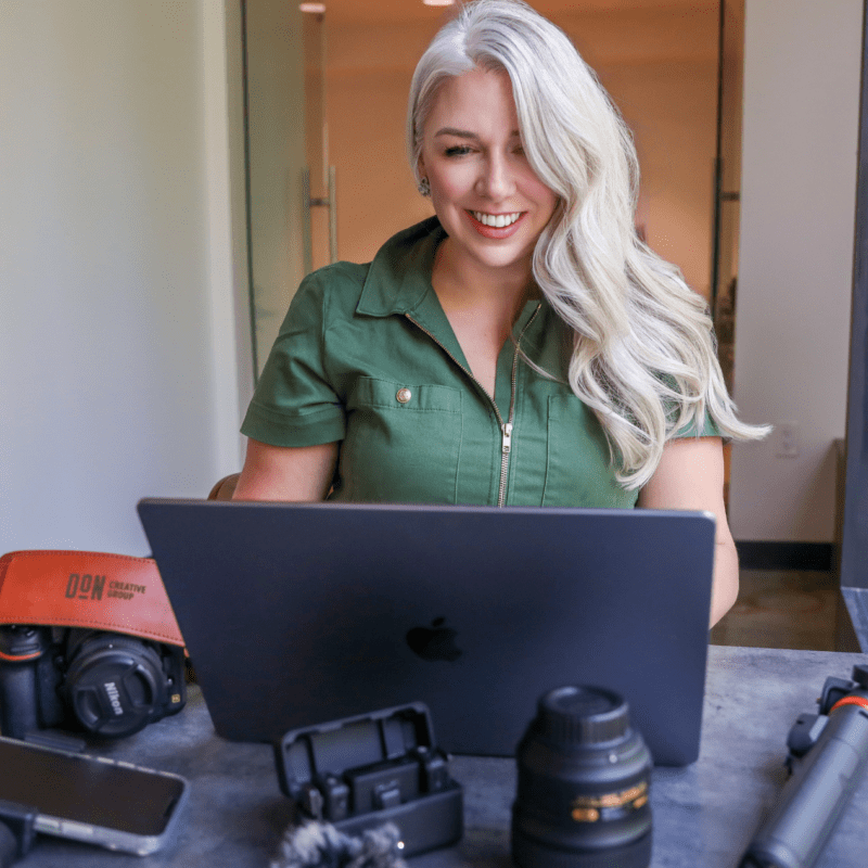 Smiling woman with long blonde hair working on a laptop at a table surrounded by camera gear, smartphone, and video equipment at Don Creative Group.