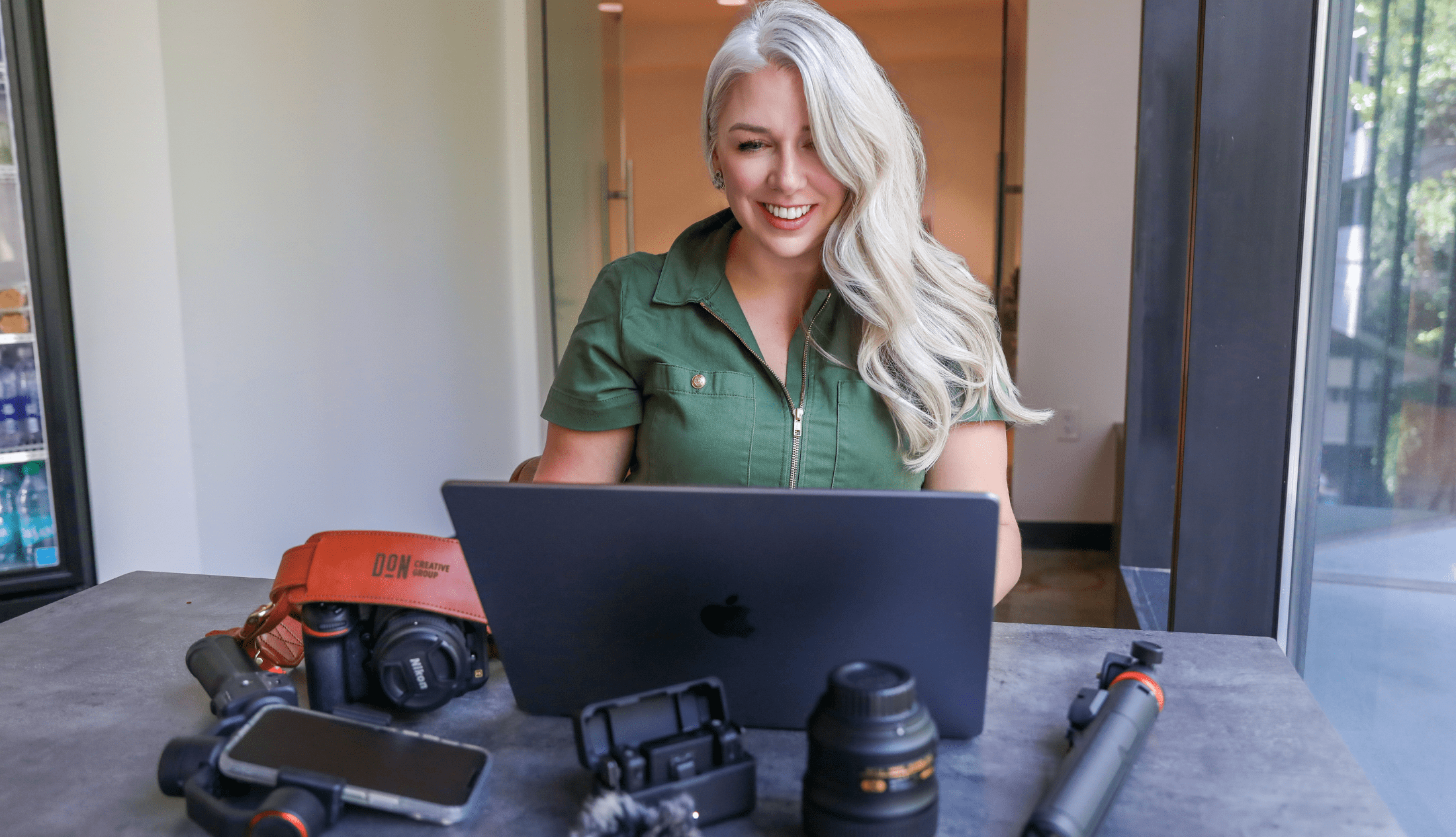 Smiling woman with long blonde hair working on a laptop at a table surrounded by camera gear, smartphone, and video equipment at Don Creative Group.