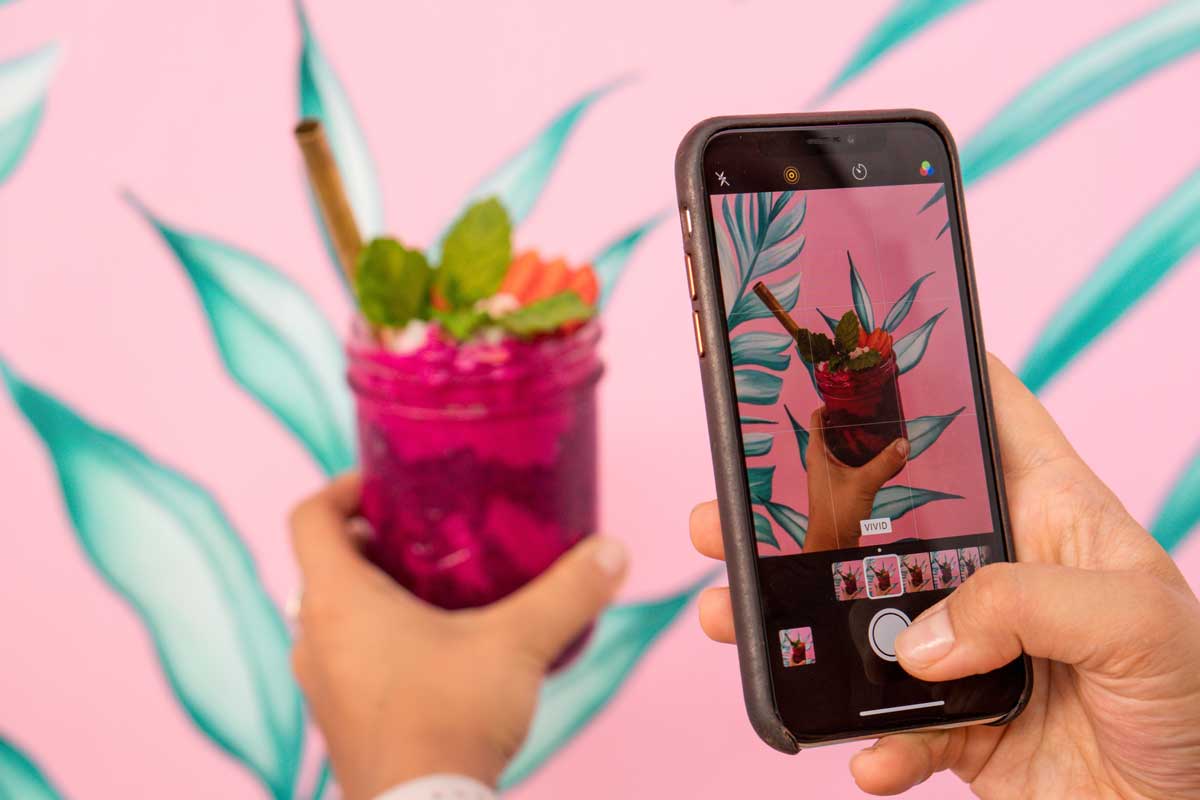 Person taking a photo of a colorful smoothie with a smartphone in front of a pink and teal Instagrammable background.