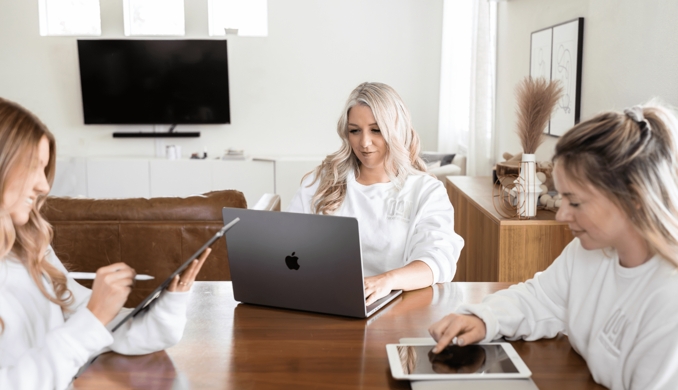 Three women from Don Creative Group collaborating at a table using laptops and tablets in a bright, modern workspace.