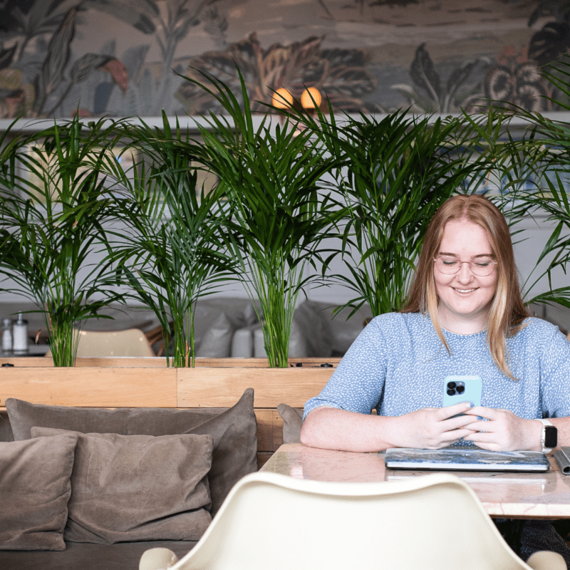 Smiling woman sitting at a marble café table with notebooks and a phone, surrounded by lush green plants and cozy modern decor.