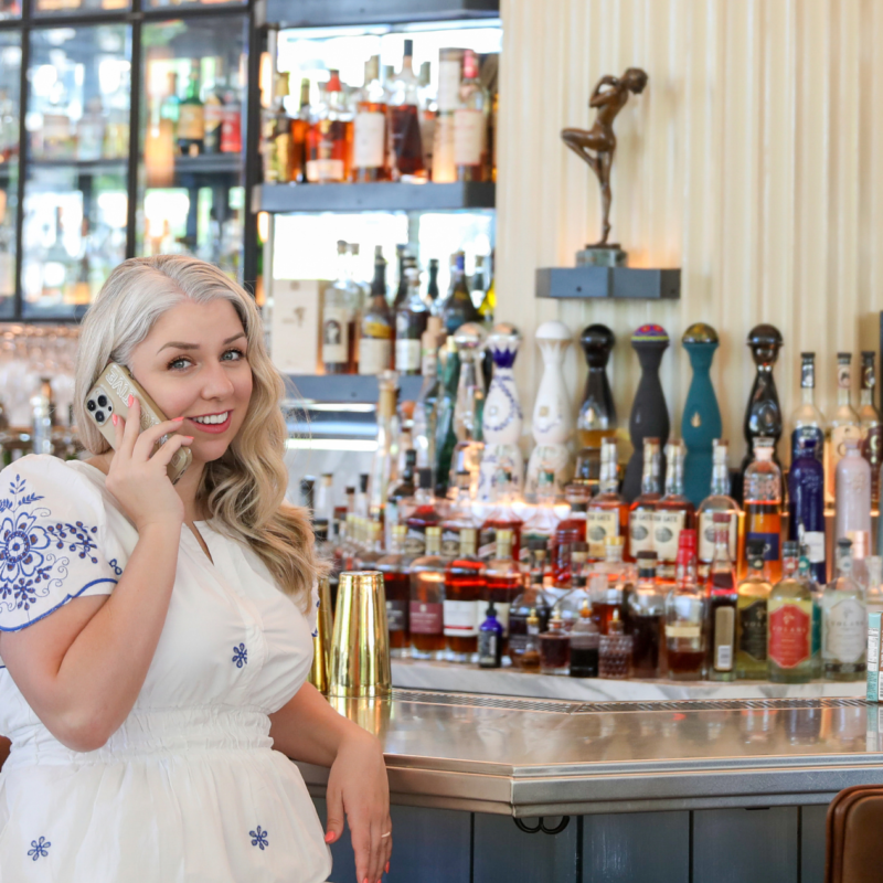 Woman in a white dress smiling while on a phone call at a stylish bar, standing in front of a well-stocked liquor display with elegant decor.
