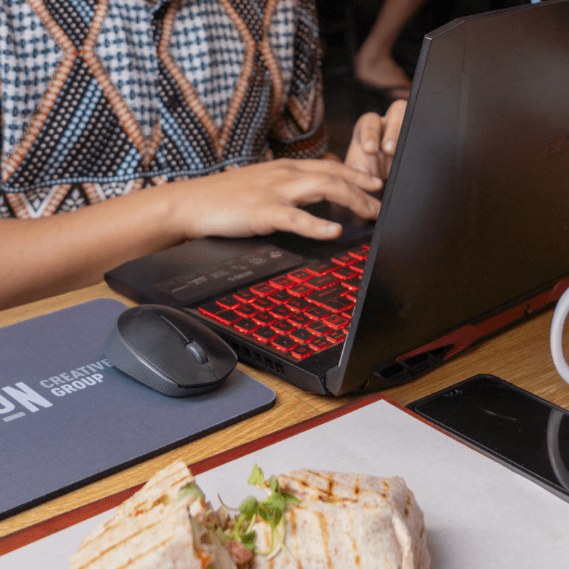 Person typing on a laptop at a wooden table with a Don Creative Group mouse pad and mug, alongside a smartphone and fresh lunch wrap.