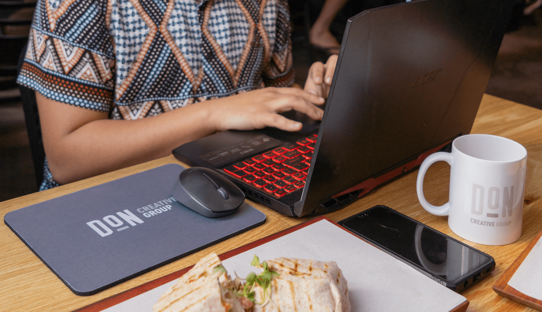 Person typing on a laptop at a wooden table with a Don Creative Group mouse pad and mug, alongside a smartphone and fresh lunch wrap.