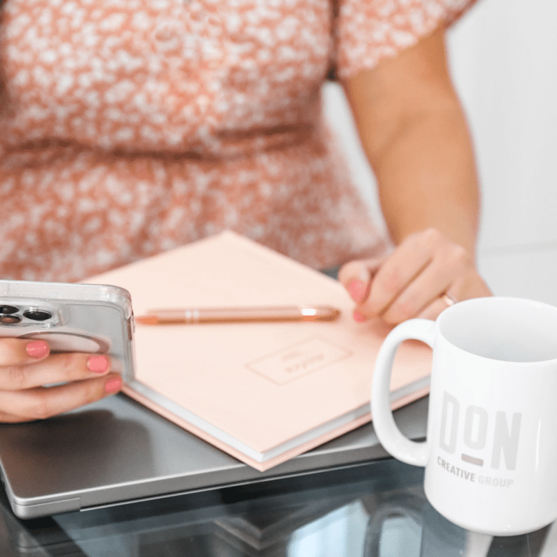 Close-up of a woman using a smartphone at a glass desk with a Don Creative Group mug, planner, and laptop—ideal for content planning or SEO strategy.