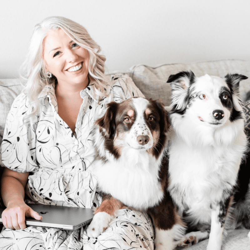 Smiling woman sitting on a couch with two Australian Shepherd dogs and a laptop, creating a cozy and joyful work-from-home atmosphere.