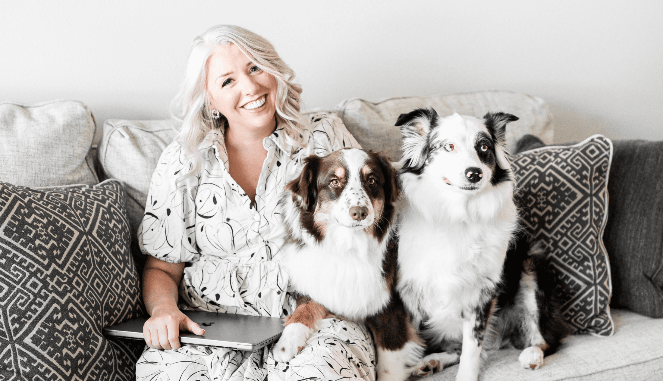 Smiling woman sitting on a couch with two Australian Shepherd dogs and a laptop, creating a cozy and joyful work-from-home atmosphere.