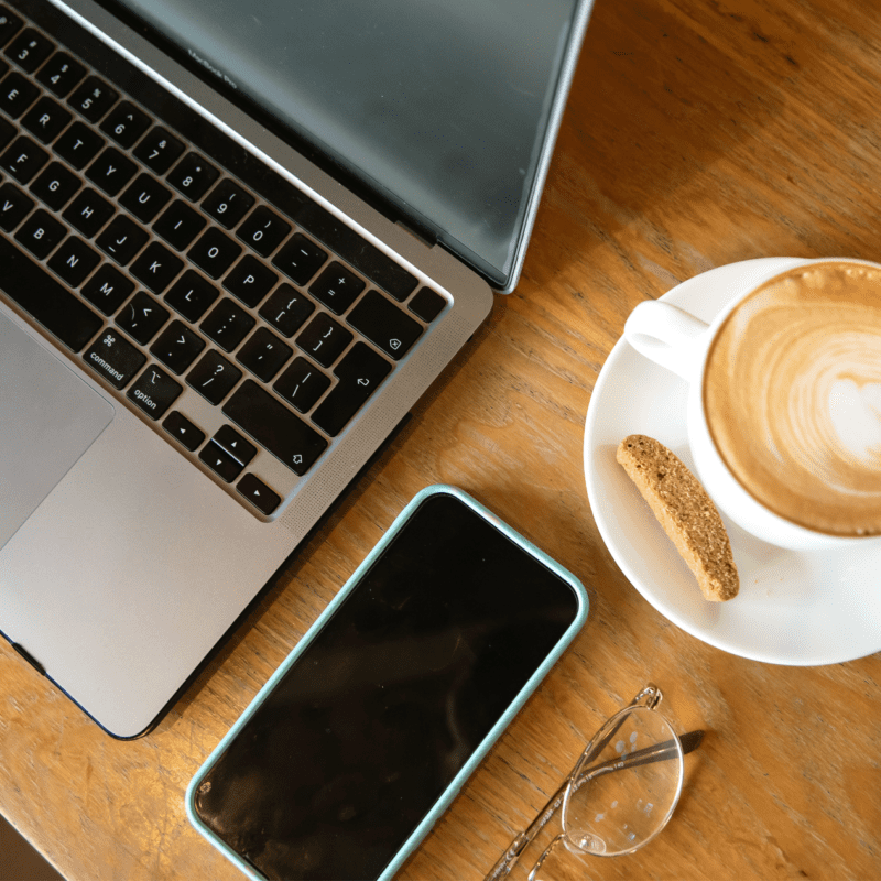 Flat lay of a workspace featuring a MacBook, smartphone, reading glasses, and a cappuccino with biscotti on a wooden table.