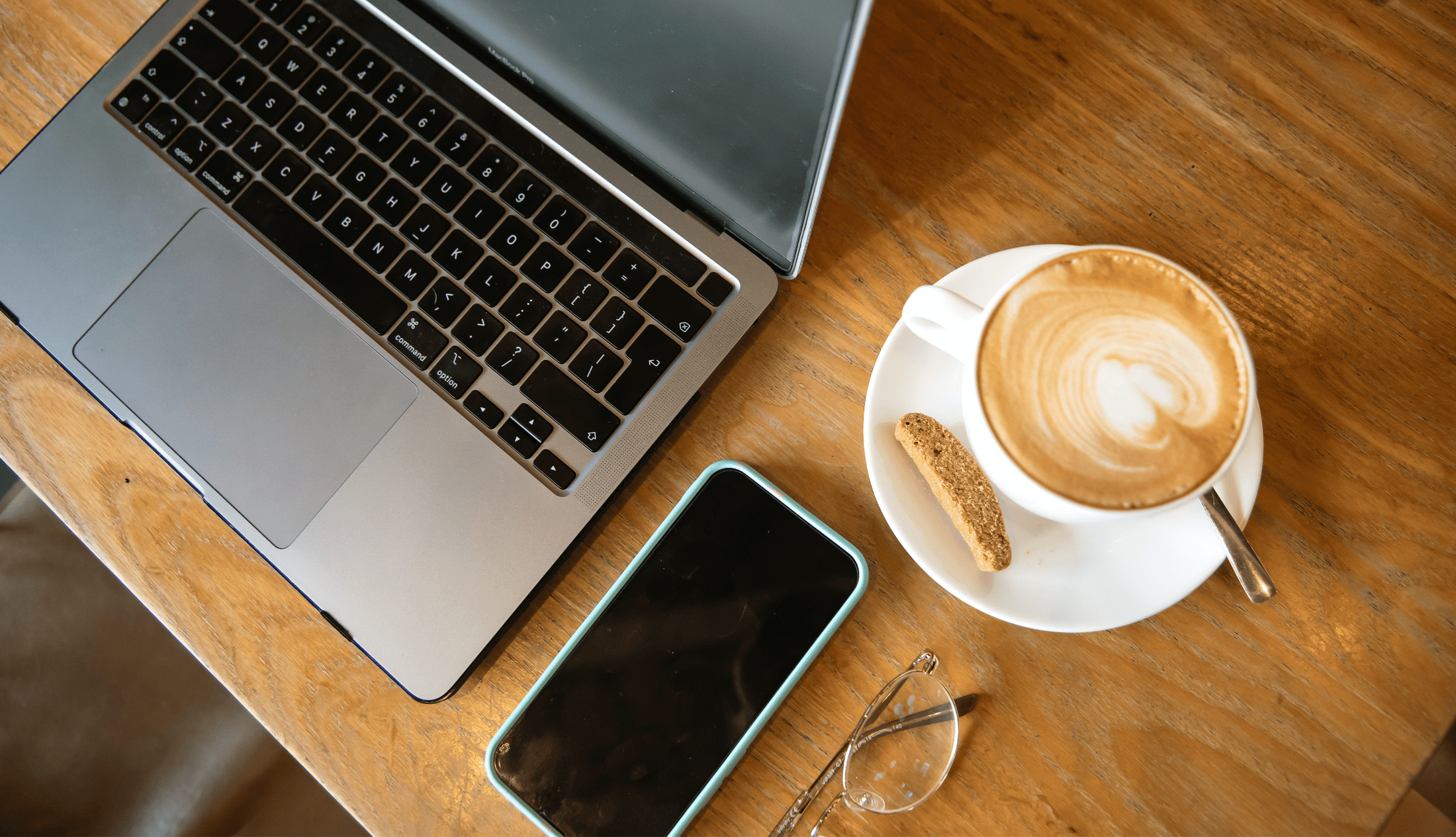 Flat lay of a workspace featuring a MacBook, smartphone, reading glasses, and a cappuccino with biscotti on a wooden table.