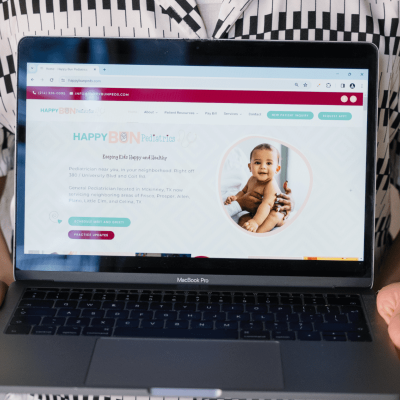Person holding a MacBook Pro displaying the Happy Bun Pediatrics website, featuring a smiling baby and service locations in Texas.