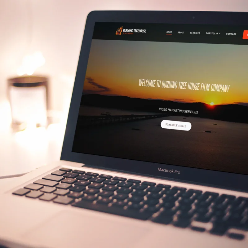Laptop displaying the homepage of Burning Tree House Film Company with a sunset background and call-to-action button for video marketing services.