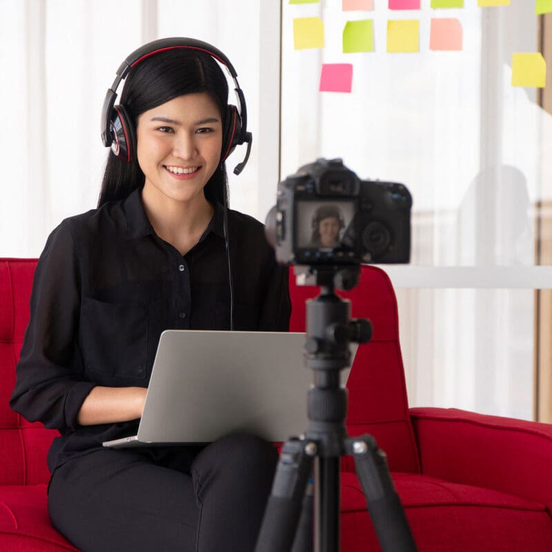 a lady sitting on a red couch with a laptop on her lap and headset on her head, smiling into a camera that is recording her for content that she will be posting for her business