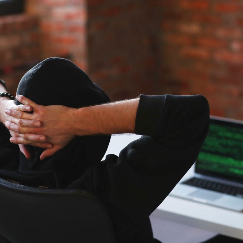 Person in a hoodie sitting at a desk with hands behind head, facing a laptop screen displaying green code symbolizing a hacker or cybersecurity threat.
