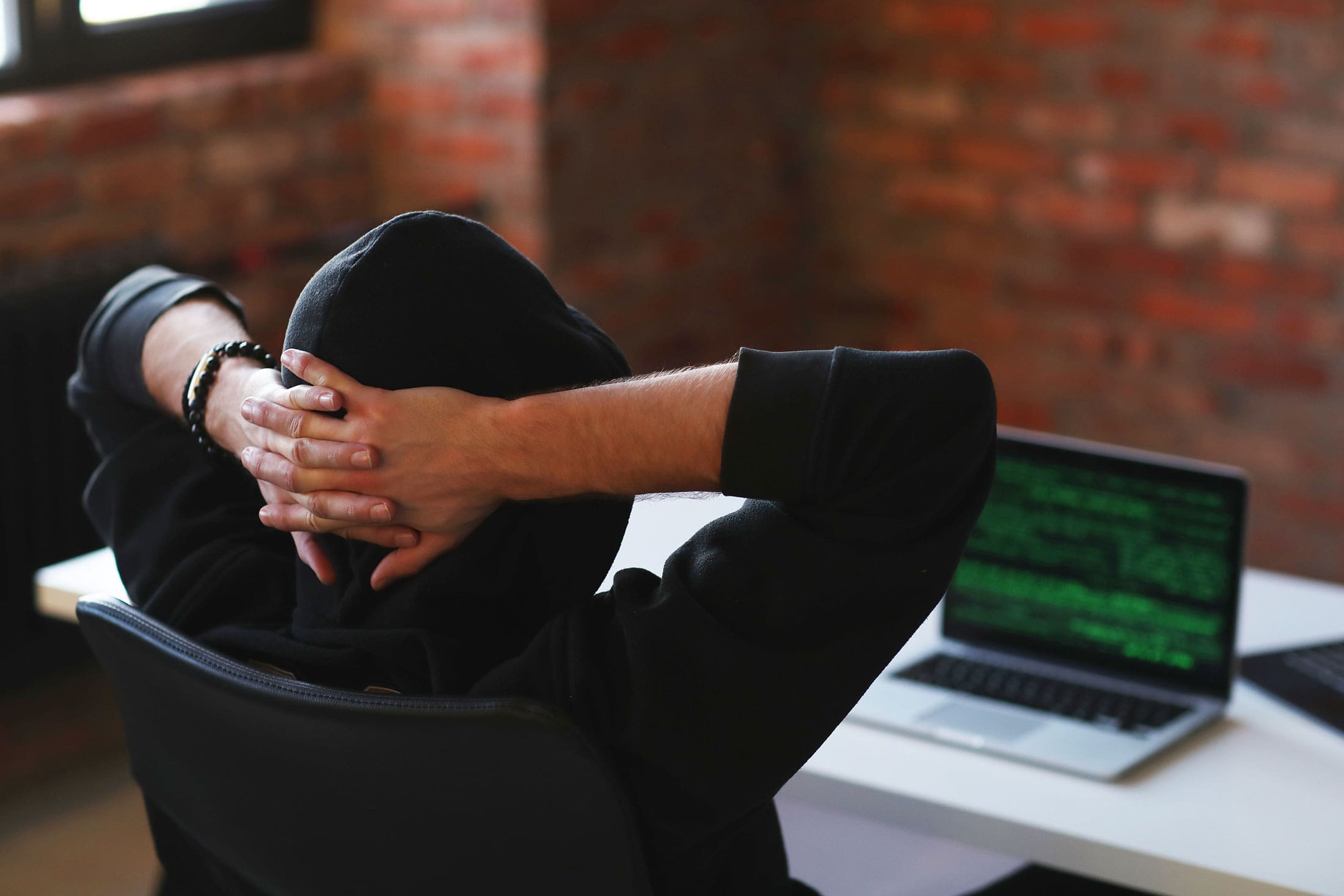Person in a hoodie sitting at a desk with hands behind head, facing a laptop screen displaying green code symbolizing a hacker or cybersecurity threat.