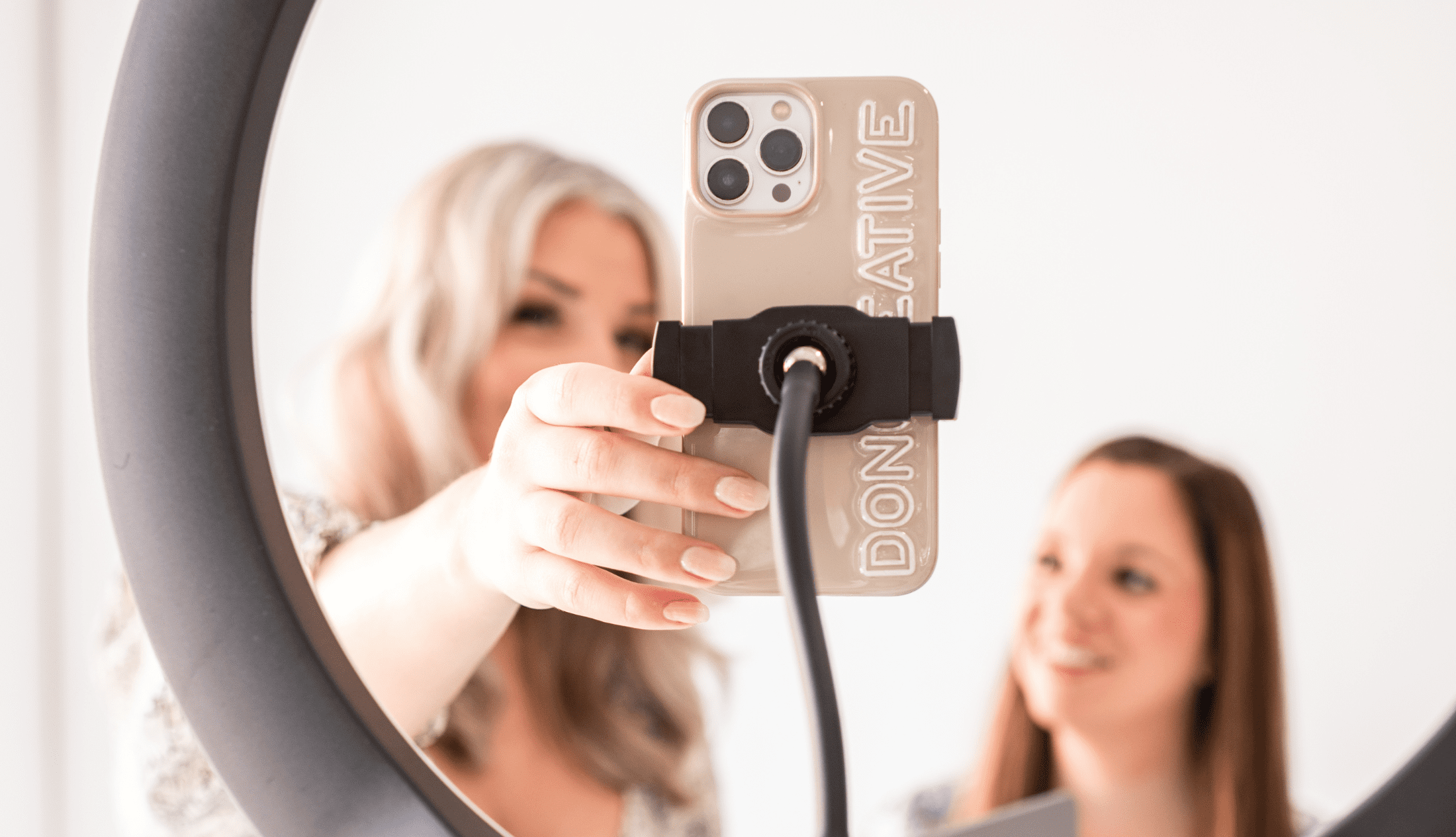 Two women setting up a smartphone in a ring light for content creation, with a custom Don Creative Group phone case in focus.