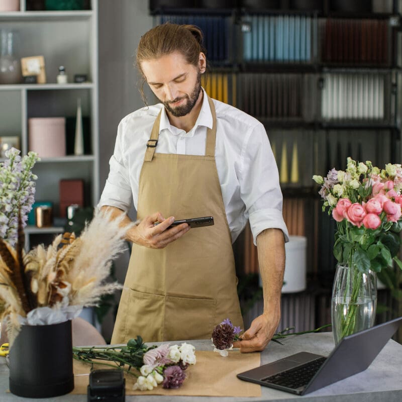 Florist in an apron using a smartphone while arranging flowers in a modern flower shop workspace