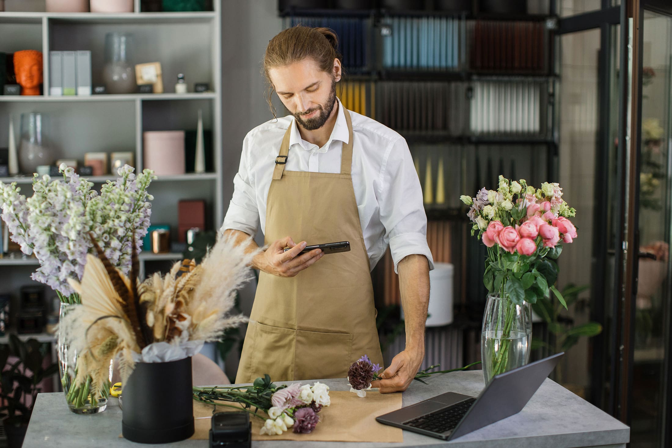 Florist in an apron using a smartphone while arranging flowers in a modern flower shop workspace