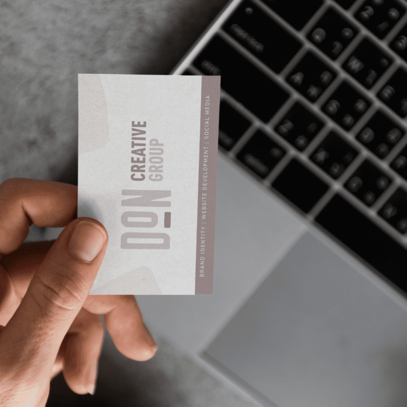 A hand holding Don Creative Group's Business card with an open laptop on the table
