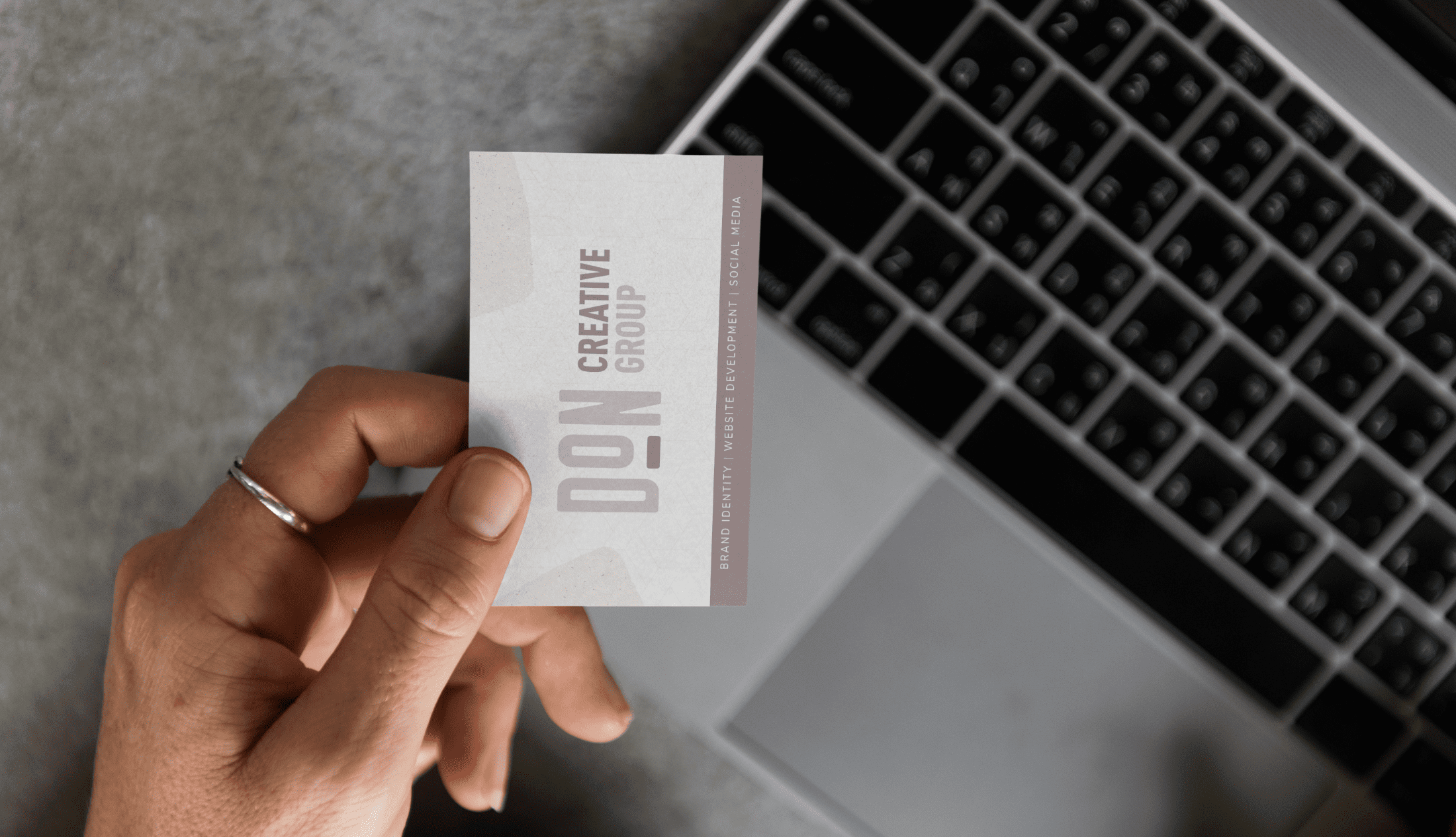 A hand holding Don Creative Group's Business card with an open laptop on the table