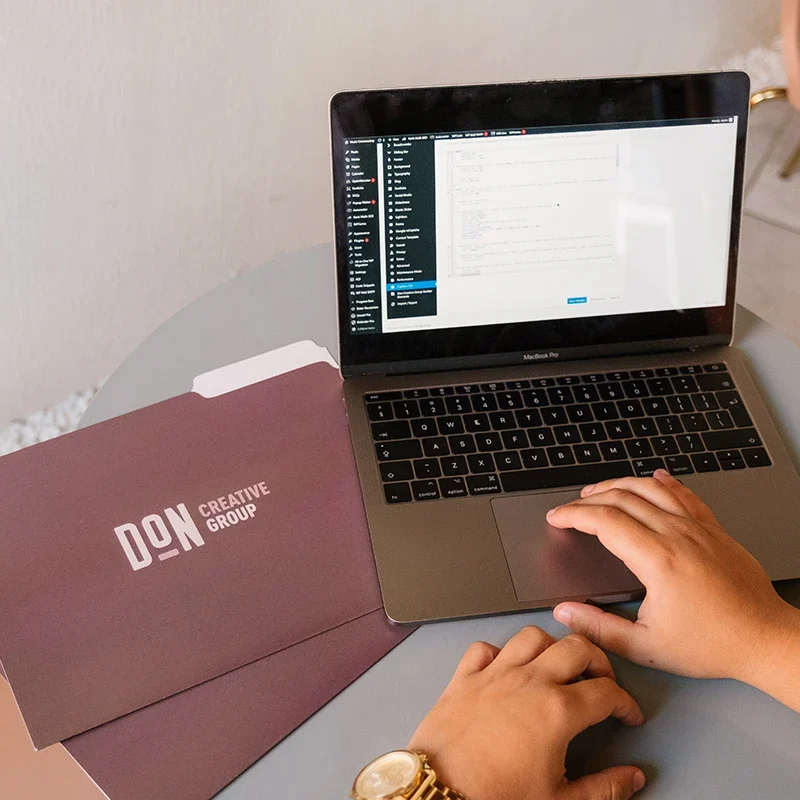 Hands working on a laptop for website development beside Don Creative Group branded folders