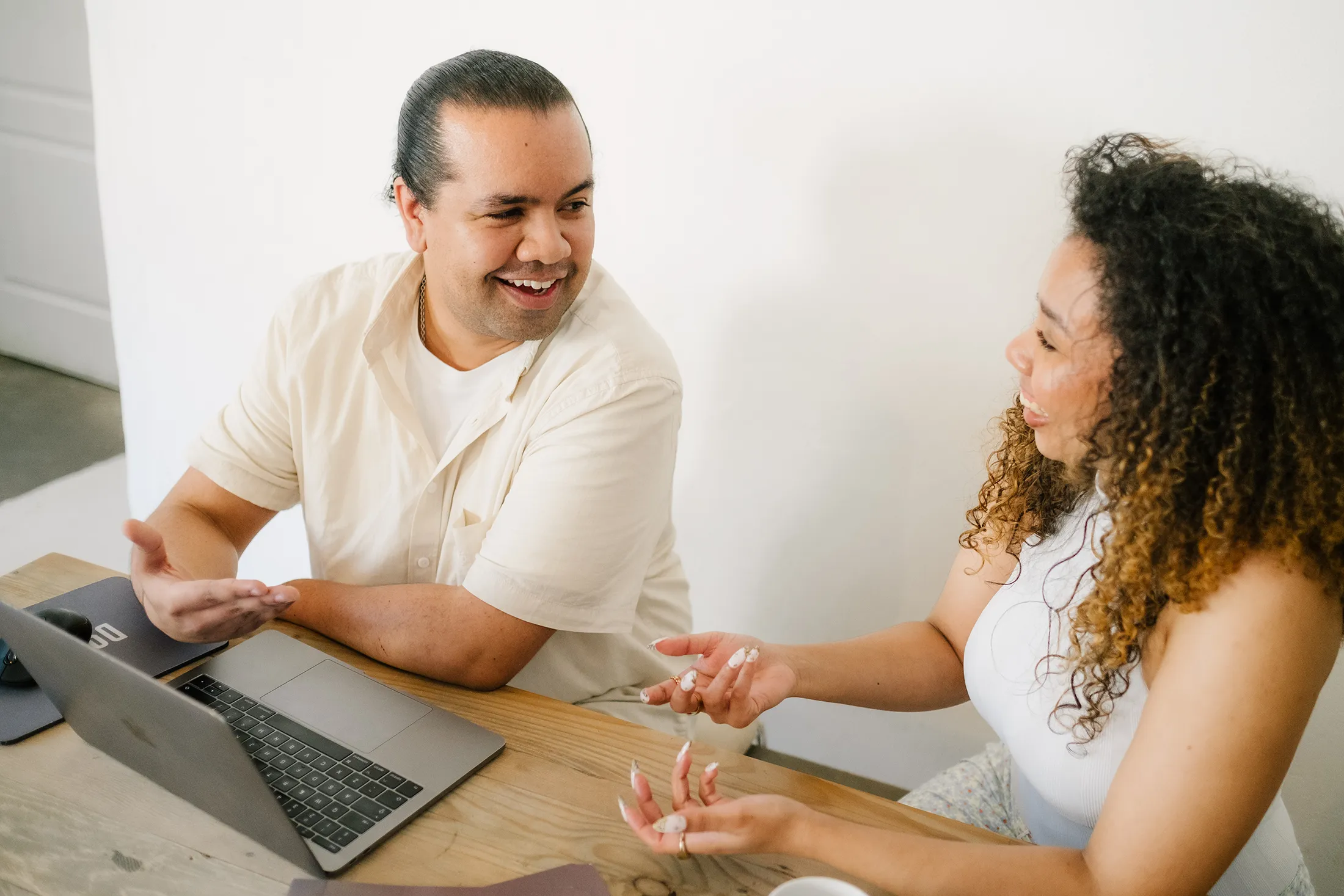 Two coworkers smiling and having a lively conversation at a desk with a laptop during a collaborative work session.