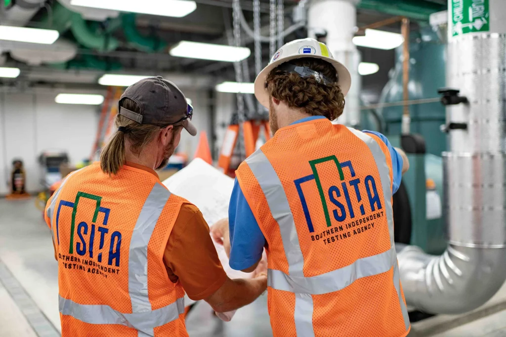 Two construction professionals wearing safety vests review plans inside a mechanical room during HVAC testing and inspection