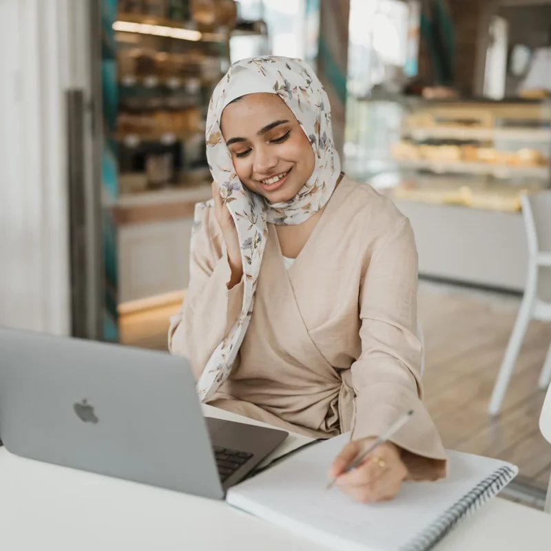 Woman working on a laptop while writing notes at a cafe, representing social media planning, content strategy development, and digital marketing workflow