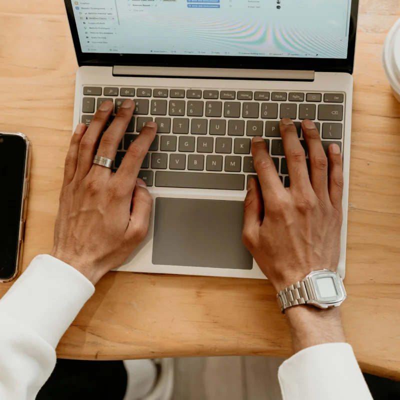 Hands typing on a laptop keyboard at a wooden desk with phone and coffee nearby