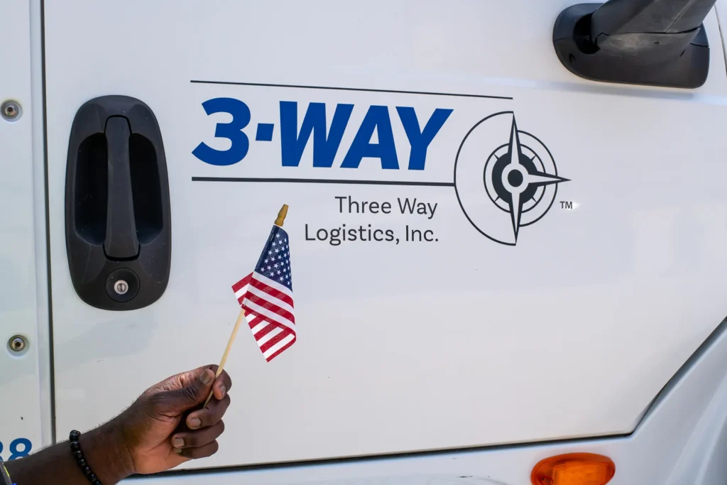 Three Way Logistics branded vehicle with American flag Hand holding a small American flag in front of a Three Way Logistics company vehicle logo